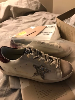 Golden Goose vita sneakers med glitter - Chic Golden Goose sneakers i vitt läder med silverglittrig stjärna på sidan och vinröda glitterdetaljer på hälen. Skorna har vita snören med svart text och en något smutsad tå för den klassiska worn looken. Perfekta för dig som gillar statement-detaljer. Storlek 40 men känns som 39.