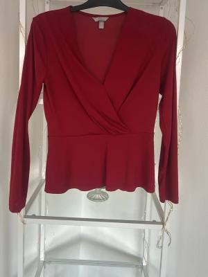Vinröd omlottopp från H&M - Snygg vinröd omlottopp från H&M med lång ärm och v-ringad skärning. Toppen har en figurnära passform och är tillverkad i ett mjukt, stretchigt material som känns bekvämt mot huden. Perfekt att styla med jeans eller kjol för en trendig look.