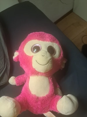 Rosa gosedjursapa med stora ögon - Söt rosa gosedjursapa med stora glittriga ögon och beige ansikte, mage, händer och fötter. Apan har ett broderat leende och mjuk svans. Perfekt som mysigt sällskap eller dekoration i rummet.