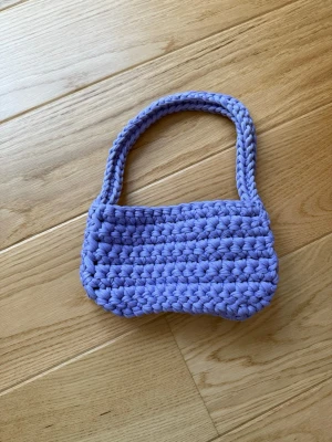 Ljuslila virkad handväska - Handgjord ljuslila handväska i virkat tjockt garn. Väskan har en mjuk, rundad form och ett matchande handtag. Perfekt för dig som gillar unika accessoarer och vill sticka ut med en färgglad detalj.