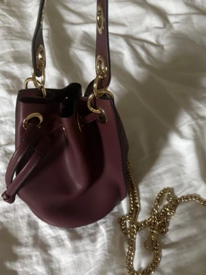 Vinröd bucket bag - Snygg vinröd bucket bag i läderimitation med bred axelrem och stora guldfärgade metalldetaljer. Väskan har en cool dragskoöppning och en lång guldkedja som axelband. Perfekt för dig som vill ha en trendig och unik handväska med statement-detaljer.