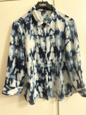 Blåvit mönstrad blus från ginatricot - Snygg blus i blått och vitt med abstrakt mönster från Gulins. Blusen har krage, knäppning framtill och långa ärmar med knapp vid ärmslut. Materialet känns lätt och luftigt, perfekt för en trendig look.