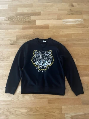 Svart Kenzo sweatshirt med tigertryck - Cool svart sweatshirt från Kenzo med ikoniskt tigertryck i vitt och gult framtill. Tröjan har rund halsringning och långa ärmar. Perfekt för dig som vill ha en statement piece i garderoben.