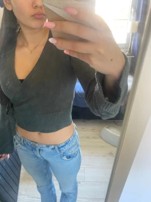 Mörkgrå omlottopp med knyt - Snygg mörkgrå omlottopp med croppad passform och knyt i sidan. Toppen har långa ärmar med bred mudd och djup v-ringning. Perfekt att styla med jeans eller kjol för en trendig look.