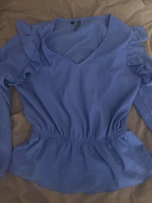 Ljusblå volangblus från Rainbow - Supersöt ljusblå blus från Rainbow med v-ringning, långa ärmar och volangdetaljer på axlarna. Blusen har resår i midjan för en snygg passform och är tillverkad i ett lätt, strukturerat tyg. Perfekt för dig som vill ha något extra till din outfit! Osäker på storlek se bild 2 men passar mig som vanligtvis är storlek S