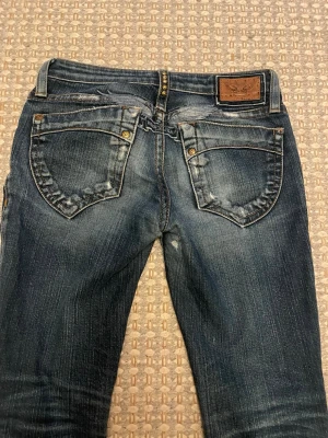 Blå jeans från Robin's Jean - Säljer ett par blå jeans från Robin's Jean med snygga slitningar och detaljerade bakfickor med nitar. Jeansen har klassisk femficksdesign, låg midja och är gjorda i denim. Märkespatch i läder baktill och coola sömmar som ger ett unikt uttryck.