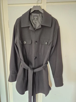 Svart vårjacka/kappa med knytband från Lindex - Jättefin svart vårjacka/kappa med knytband i midjan från Lindex. Storlek M men skulle säga att den är mer som en M/L. I nyskick!