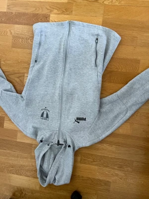 Grå hoodie Malmö FF x Puma - Snygg ljusgrå hoodie från Puma i samarbete med Malmö FF. Tröjan har dragkedja framtill, två fickor med dragkedja och tryck med både Puma-logga och Malmö FF-emblem på bröstet. Tillverkad i mjukt sweatshirtmaterial och har justerbar huva med snören.