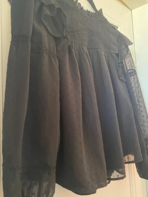 Svart volangblus med lång ärm ifrån Gina young❣️❣️170 men känns som M🤍 - Snygg svart blus med volangdetaljer och smockad överdel. Blusen har transparenta långärmar med diskret prickmönster och spetsdetaljer vid ärmslut. Luftig passform och rundad halsringning. Perfekt för dig som gillar romantisk och trendig stil. 