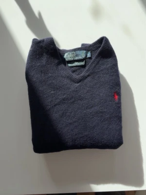 Mörkblå v-ringad tröja Polo Ralph Lauren - Klassisk mörkblå v-ringad tröja från Polo Ralph Lauren med den ikoniska röda logotypen broderad på bröstet. Tröjan har långa ärmar och ribbade muddar vid ärmslut och nederkant. Perfekt för lager på lager och stilrena outfits. Den är lite smått krymt i tvätten då det är ull men sitter superbra som S och finns inga andra tecken på skavanker 😊