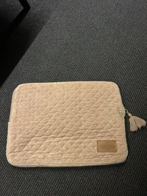 datorväska med tofs - Säljer en beige quiltad datorväska med dragkedja och dekorativ tofs. Väskan har ett mjukt sammetstyg och en liten läderpatch med logga på framsidan. Perfekt för att skydda din laptop med stil.