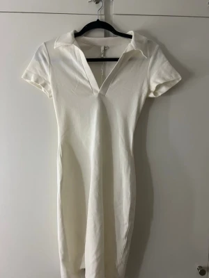 Vit kortklänning med krage - Stilren vit kortklänning med krage och v-ringad hals. Klänningen har korta ärmar och en figurnära passform som ger en snygg siluett. Perfekt för dig som gillar minimalistisk och modern stil.