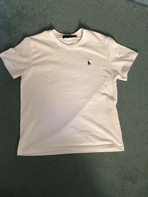 Vit t-shirt från Polo Ralph Lauren - Klassisk vit t-shirt från Polo Ralph Lauren med rund hals och korta ärmar. Diskret broderad logga i svart på bröstet. Tillverkad i mjuk bomull för en skön och avslappnad look.
