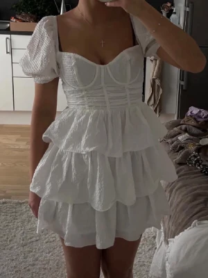 Vit volangklänning med puffärm - Supersöt vit kortklänning med flera lager volanger och puffärmar. Klänningen har markerad byst, smockad struktur och fyrkantig urringning. Perfekt för dig som vill ha en romantisk och trendig look. Materialet känns lätt och luftigt.