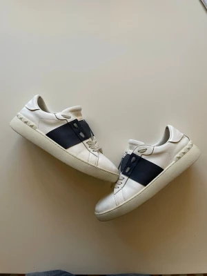 Valentino opens marinblå - Snygga vita sneakers från Valentino med bred svart rem över sidan och nitar längs hälen. Tillverkade i läder med klassisk låg siluett och vita snören. Perfekta för dig som vill ha en stilren men ändå unik look.
