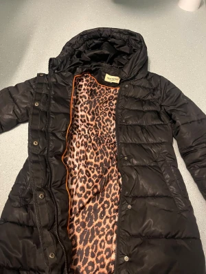 Svart lång dunjacka Roco Baroco - Snygg svart lång dunjacka från Roco Baroco med leopardmönstrat foder och orange kantdetalj. Jackan har dragkedja och tryckknappar framtill, perfekt för kalla vinterdagar. Stilsäker och varm design som sticker ut med sitt coola innerfoder.