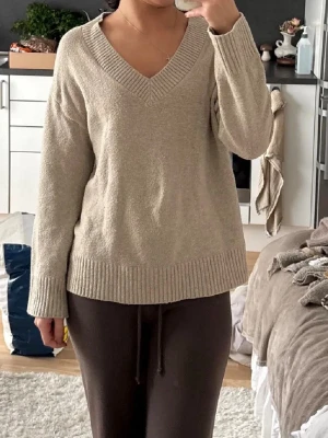 Beige stickad tröja med v-ringning - Mysig beige stickad tröja med djup v-ringning och breda ribbade muddar vid ärmslut och nederkant. Tröjan har en avslappnad passform och är perfekt för lager-på-lager. Materialet känns mjukt och bekvämt mot huden.
