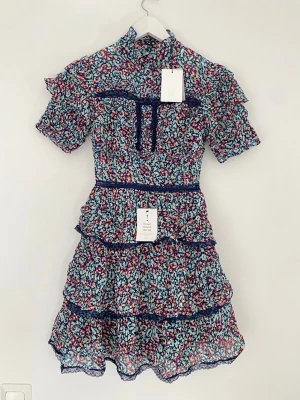 Blommig volangklänning från By Malina - Supersöt blommig klänning från By Malina med volanger, hög krage och spetsdetaljer. Klänningen har korta ärmar och ett färgstarkt mönster i blått, rosa och vitt. Perfekt för dig som gillar romantisk och lekfull stil.