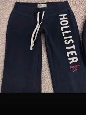Mågmidjade mjukisbyxor Hollister - Mörkblå lågmidjade mjukisbyxor från Hollister med vit dragsko i midjan och stort vitt tryck längs benet där det står 'HOLLISTER' samt röd text 'Newport Bch'. Sköna och avslappnade med raka ben och mjukt material.