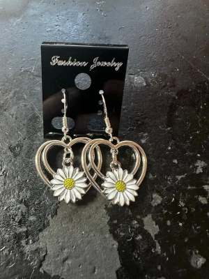 Hjärnformade örhängen med prästkrage - Säljer ett par örhängen i silverfärgad metall med hjärtformad design och hängande prästkrageblommor i vitt och gult. Perfekta för dig som vill ha en söt och unik accessoar som sticker ut. 