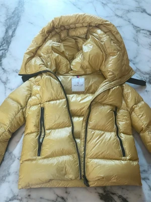 Gul dunjacka från Moncler begagnad  - Snygg gul dunjacka från Moncler med glansig finish och stor huva. Jackan har två svarta dragkedjeförsedda fickor framtill och klassisk Moncler-logga på ärmen. Perfekt för kalla vinterdagar och riktigt trendig look.