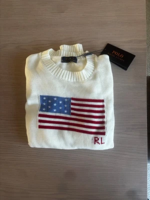 Vit stickad tröja Polo Ralph Lauren - Vit stickad tröja från Polo Ralph Lauren med amerikansk flagga i rött, vitt och blått på bröstet samt RL-broderi. Tröjan har rund halsringning och ribbade muddar vid ärmslut och nederkant. Perfekt för dig som gillar klassisk preppy stil.