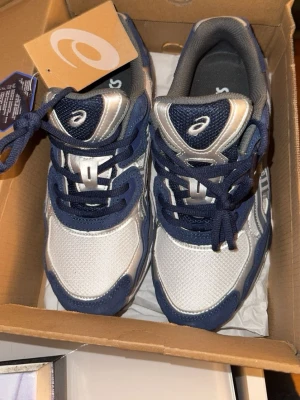 Asics vita och blå sneakers - Snygga sneakers från Asics i vitt och marinblått med silverdetaljer. Skorna har en klassisk låg siluett, mesh- och syntetmaterial samt tydliga loggor på plösen och sidan. Perfekta för dig som gillar sportig och trendig stil.