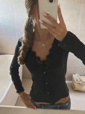 Svart långärmad topp med spets - Snygg svart långärmad topp med knappar framtill och v-ringad urringning dekorerad med spets. Figurnära passform som framhäver siluetten. Perfekt att styla med jeans eller kjol för en trendig look.