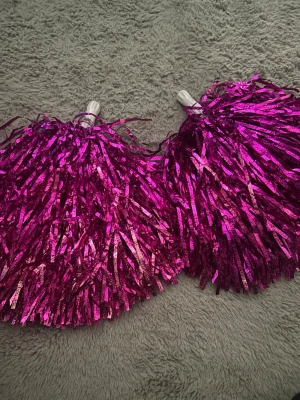 Rosa metallic pompoms cosplay - Två stycken glittriga rosa pompoms med vita handtag, perfekta för maskerad, cheerleading eller cosplay. Tillverkade i plast med metallicfinish som ger extra effekt när du rör på dem.