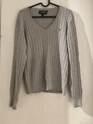 Grå kabelstickad tröja från GANT - Snygg grå kabelstickad tröja från GANT med V-ringning och diskret logga broderad på bröstet. Tröjan har långa ärmar och ribbade muddar vid ärmslut och nederkant. Perfekt för lager på lager och enkel att matcha till olika outfits.