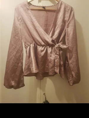 Ljuslila omlottblus med knytband - Snygg ljuslila omlottblus i glansigt material med långa ärmar och volang nedtill. Blusen har ett knytband i midjan som ger en feminin siluett och djup v-ringning. Perfekt för dig som gillar trendiga och eleganta plagg. Storlek: M