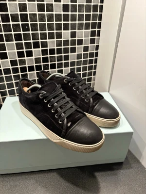 Lanvin cap toe  - Stilrena svarta sneakers med ovandel i mocka och tåparti i slätt läder. Skorna har svarta skosnören, metallöljetter och vit sula med diskret randstruktur. Perfekta för dig som gillar en clean och klassisk look.
