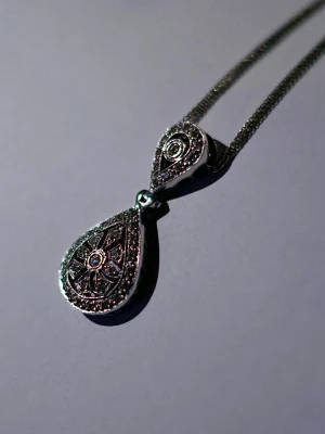 Silverfärgat halsband med droppformad berlock - Unikt halsband i silverfärg med en droppformad berlock dekorerad med små gnistrande stenar och detaljerat mönster. Kedjan är tunn och elegant, perfekt för att lyfta vilken outfit som helst. Berlocken har en vintageinspirerad design med fina ornament.