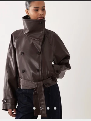 Brun oversized skinnjacka med bälte - Snygg mörkbrun skinnjacka med dubbelknäppning, bred krage och bälte i midjan. Jackan har en oversized passform och långa ärmar med knappdetaljer vid ärmslut. Perfekt för dig som vill ha en trendig och edgy look.