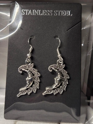 Måneformade örhängen i stål - Unika örhängen i rostfritt stål med måneform och ansikte i profil. Dekorerade med detaljerade prickar och bladliknande mönster runt månen. Silverfärgade och hängande modell som ger en cool och mystisk vibe till din stil. Hänget är ca 2 cm.