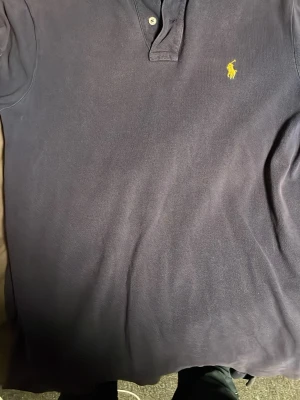 Mörkblå pikétröja från Ralph Lauren - Klassisk mörkblå pikétröja från Ralph Lauren med gul broderad logga på bröstet. Tröjan har korta ärmar, krage och knappar vid halsen. Tillverkad i bomull för en bekväm och stilren look.