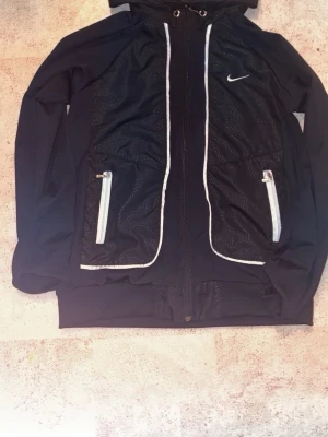 Nike tröja - Snygg svart tröja från Nike med vita dragkedjor och detaljer. Jackan har huva med dragsko, två fickor framtill och diskret Nike-logga på bröstet. Perfekt för en sportig och avslappnad stil.