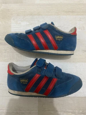 Adidas sneakers - Säljer ett par klassiska Adidas  sneakers i blå mocka och mesh med röda detaljer och kardborreband. Skorna har de ikoniska tre ränderna på sidan och guldfärgad logga. Låg modell med vit och blå sula. Perfekta för dig som gillar retrostil.