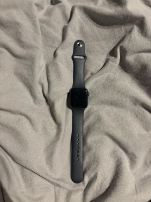 Svart Apple Watch med silikonarmband - Snygg svart Apple Watch med rektangulär skärm och matchande svart silikonarmband. Klockan har en modern och stilren design med touchskärm och justerbart band som passar de flesta handleder. Perfekt för dig som vill ha koll på notiser, träning och hälsa direkt på handleden.