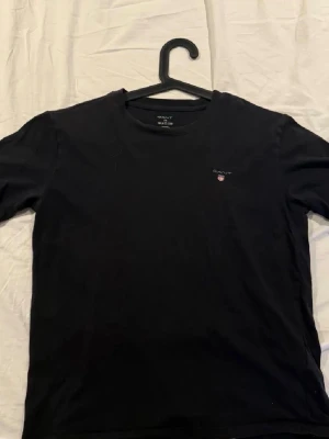 svart t-shirt från GANT - Snygg marinblå t-shirt från GANT med klassisk rund hals och liten logga med sköld på bröstet. Tillverkad i mjuk bomull för skön känsla och enkel passform. Perfekt basplagg till garderoben.