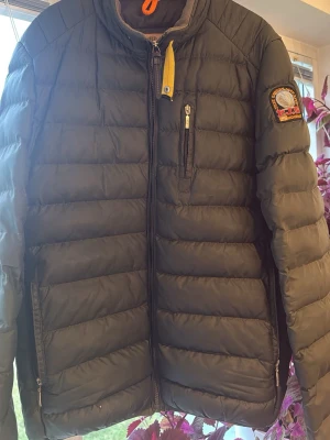 Svart dunjacka från Parajumpers - Snygg svart dunjacka från Parajumpers med gul detalj vid halsen och patch på ärmen. Jackan har en bröstficka med dragkedja, två sidofickor och quiltad design. Perfekt för kalla vinterdagar men passar även till höst och tidig vår. riktigt stilren look. Passar M-L. Pris går diskutera 