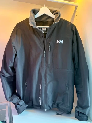 Svart Helly Hansen vindjacka - Snygg svart vindjacka från Helly Hansen med vit logga på bröstet och längs dragkedjan. Jackan har hög krage med grå insida. Storlek S