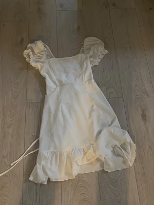 Vit kortklänning med volang och puffärm - Supersöt vit kortklänning med puffärmar och volangkant nertill. Klänningen har snörning i ryggen och markerad midja för en feminin siluett. Perfekt för sommarens alla tillfällen! Säljer pga kommer inte till användning, är storlek M men passar S med. Riktigt fin klänning till student, skolavslutning, konfirmation eller bara sommaren 💕