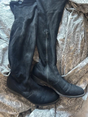 Svarta overknee stövlar i mockaimitation - Säljer ett par svarta overknee stövlar i mjuk mockaimitation. De har låg klack, rund tå och dragkedja på insidan för enkel påtagning. Perfekta till höstens och vinterns outfits för en snygg och trendig look.