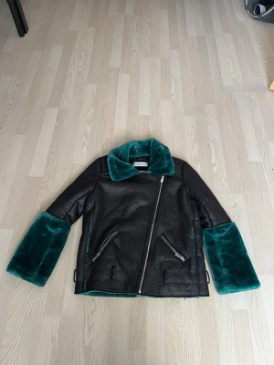 Svart pälsjacka med grön fuskpäls - Svart pälsjacka med grön fuskpäls på krage, ärmslut och insida. Jackan har snedställd dragkedja, två dragkedjefickor och detaljer i skinnimitation. Passar unisex hade jag sagt