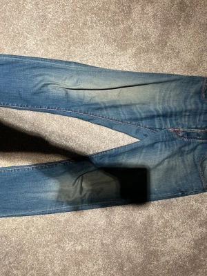 Blå jeans med röda detaljer jocob cohën  - Säljer ett par blå jeans med snyggt slitna partier och röda sömmar på bakfickorna. Klassisk femficksmodell med normal passform och raka ben. Jeansen har en röd patch bak i midjan och är tillverkade i denim.