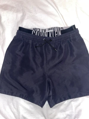 Marinblå badbyxor Calvin Klein - Snygga marinblå badbyxor från Calvin Klein med resårmidja och snörning framtill. Klassisk passform med ficka bak och tydlig logga på linningen. Perfekta för stranden eller poolen, tillverkade i snabbtorkande material.
