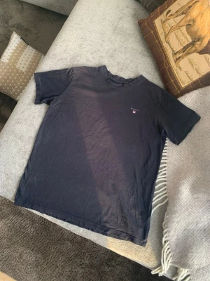 Mörkblå t-shirt från Gant - Säljer en klassisk mörkblå t-shirt från Gant med liten broderad logga på bröstet. T-shirten har rund hals och korta ärmar, perfekt för en enkel och stilren look. Tillverkad i mjuk bomull som känns skön mot huden, går att tvätta upp till 40 grader. 