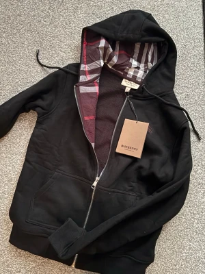 Svart hoodie från Burberry med dragkedja - Snygg svart hoodie från Burberry med dragkedja och klassiskt rutigt foder i huvan. Hoodien har fickor framtill, ribbade muddar och justerbar dragsko i huvan. Perfekt för dig som vill ha en stilren och exklusiv look med en twist av Burberrys ikoniska mönster.