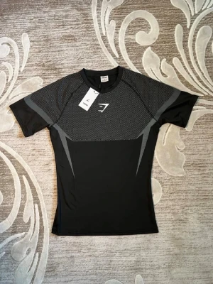 Svart träningströja från Gymshark - Snygg svart träningströja från Gymshark med gråa detaljer och logga på bröstet. Tillverkad i ett stretchigt och snabbtorkande material som passar perfekt för gymmet eller löpning. Kortärmad och med åtsittande passform för en sportig look.